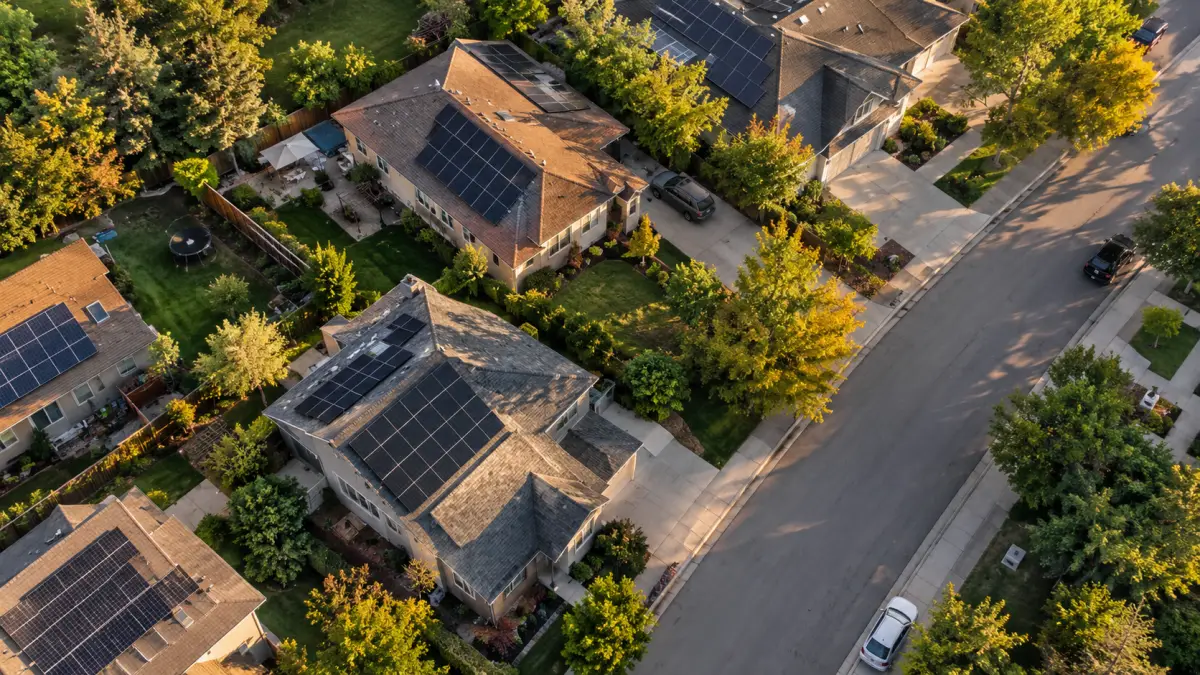 Aerial view of a suburban street with rooftop solar arrays on several homes in late-afternoon light